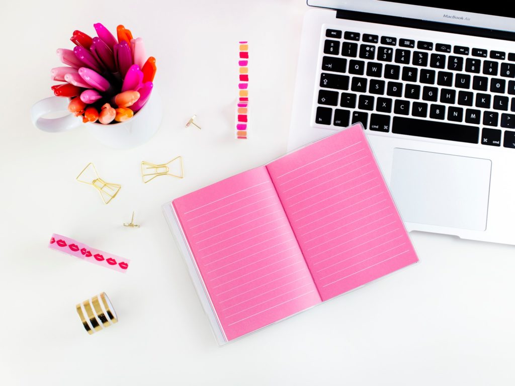 Image of a set of pens, a notebook and a laptop. Tools that people might use to write a blog post. The dominant colours are pink and white. 
